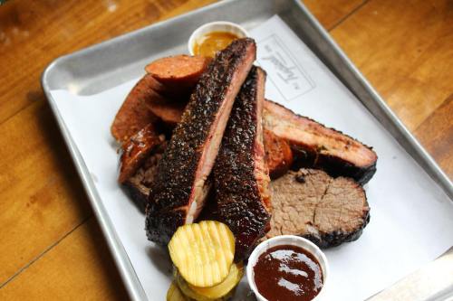 A platter of smoked meats including ribs, sausage, and brisket, served with pickles and dipping sauces.