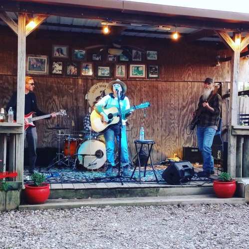 A live band performs on a wooden stage with instruments, surrounded by potted plants and framed photos on the wall.