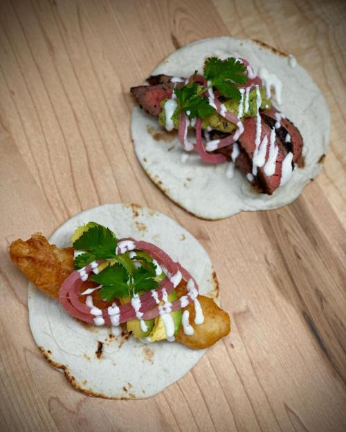 Two tacos on a wooden surface: one with grilled meat and the other with fried fish, topped with onions, cilantro, and sauce.