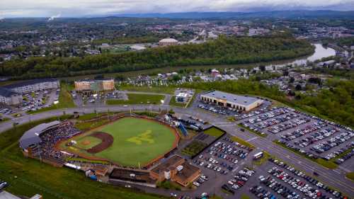 Aerial view of a baseball field surrounded by parking lots, buildings, and a river in the background.