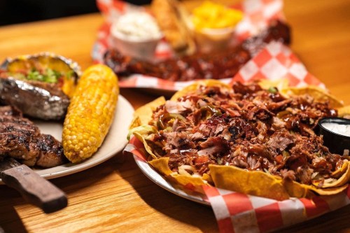 A plate of pulled pork nachos with toppings, alongside a baked potato, corn on the cob, and ribs on a wooden table.