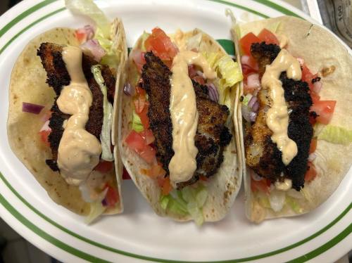 Three tacos filled with grilled fish, lettuce, tomatoes, onions, and drizzled with a creamy sauce on a white plate.
