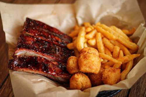 A basket of BBQ ribs, golden fries, and crispy corn fritters on parchment paper.