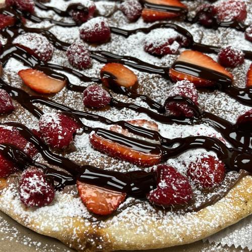 A dessert pizza topped with strawberries, raspberries, powdered sugar, and drizzled with chocolate sauce.