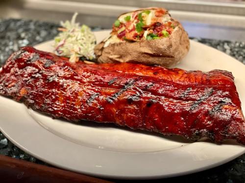 A plate featuring a large, glazed barbecue rib, a baked potato topped with bacon, and a side of coleslaw.