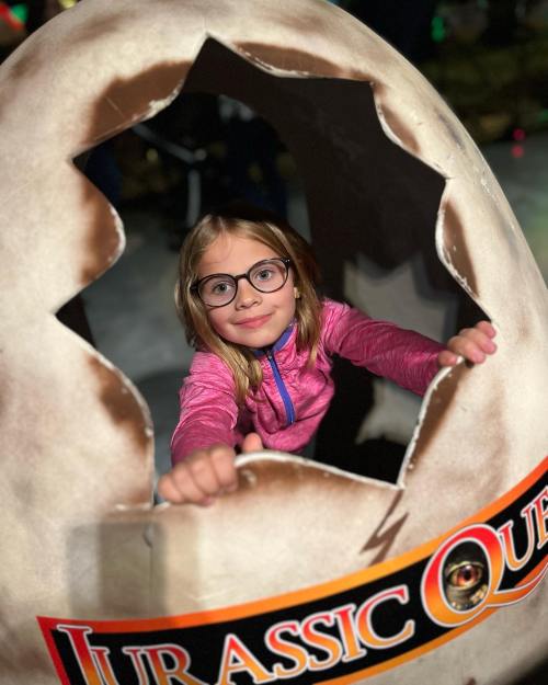A young girl with glasses peeks out from a large dinosaur egg prop at a Jurassic-themed event.