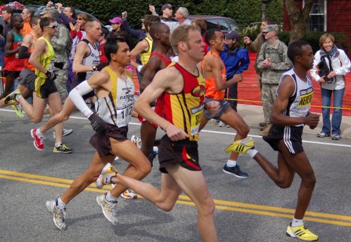 A group of male runners in athletic gear competing in a race, with spectators cheering in the background.