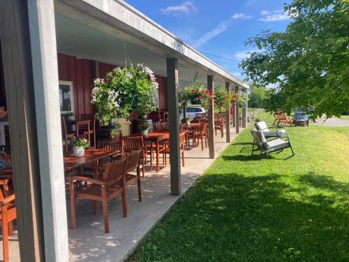 Outdoor seating area with wooden tables and chairs, surrounded by hanging plants and a grassy lawn.