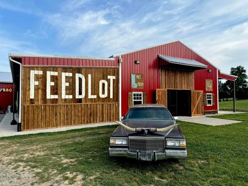 A rustic barn with "FEEDLOT" signage, featuring a classic car with horns parked in front on a grassy area.