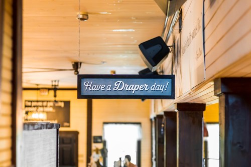 A sign hanging in a restaurant reads, "Have a Draper day!" against a warm, wooden interior backdrop.