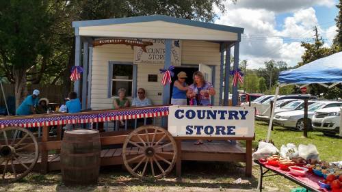 A quaint country store with people celebrating outside, decorated with red, white, and blue bunting.