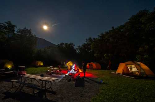 A campsite at night with tents, a picnic table, and people gathered around a fire under a bright moon.