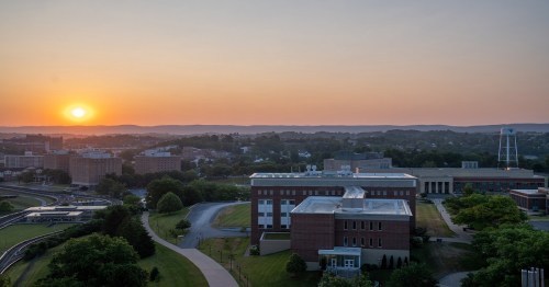 Sunrise over a campus with buildings and greenery, set against rolling hills in the background.