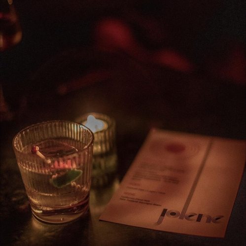 A dimly lit table with a cocktail, a lit candle, and a menu for "Jolene" in a cozy setting.