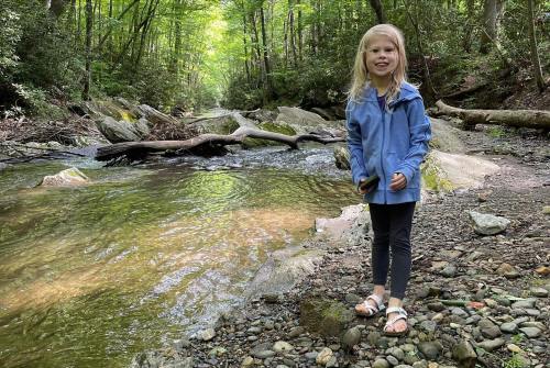 A young girl stands by a rocky stream in a forest, smiling and wearing a blue jacket and sandals.