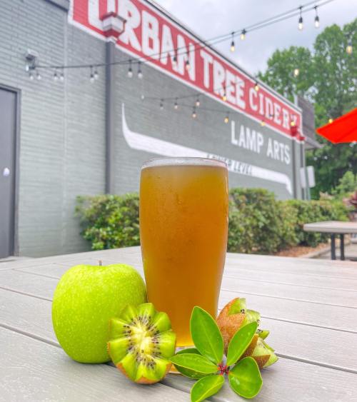 A glass of cider sits on a table with a green apple and other fruits, against a backdrop of a cider house.