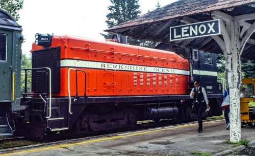 A bright orange locomotive labeled "Berkshire Scenic" at a train station with a sign reading "Lenox." A conductor stands nearby.