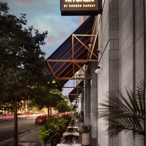 Outdoor dining area of a restaurant with elegant tables, greenery, and a modern building facade at dusk.