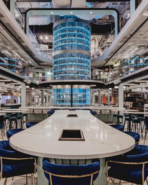 Modern dining space with a large central bar, blue lighting, and sleek white tables surrounded by stylish chairs.