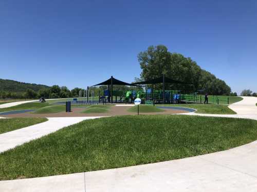 A playground with green equipment under a shaded area, surrounded by grass and a paved path on a sunny day.