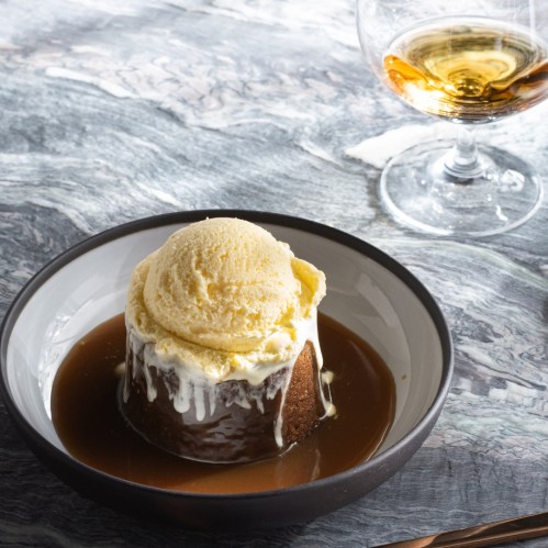 A warm dessert with ice cream on top, served in a bowl with caramel sauce, alongside a glass of amber liquid.