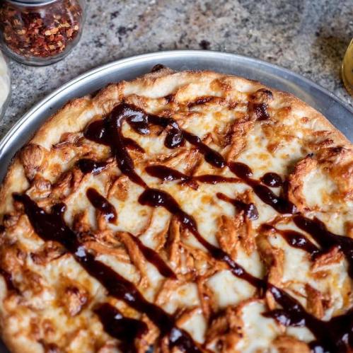 A close-up of a pizza topped with shredded chicken and drizzled with barbecue sauce on a metal tray.