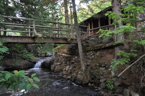A wooden bridge spans a stream, with a cabin nestled among trees and rocks in a serene forest setting.