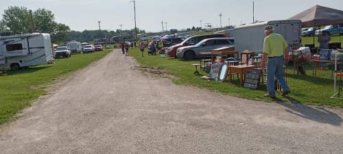 A gravel path lined with vendors and parked vehicles at an outdoor market on a sunny day.