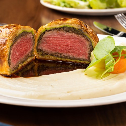 A plated beef Wellington, sliced to reveal pink meat, served with creamy puree and garnished with vegetables.