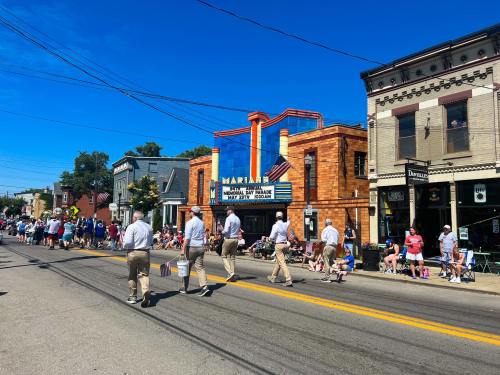 A parade scene with people walking along a street lined with spectators and colorful buildings under a clear blue sky.