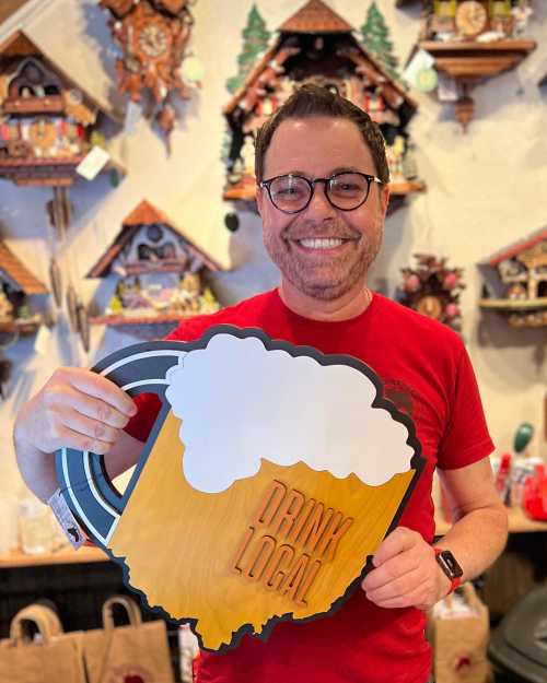 A smiling man in a red shirt holds a sign shaped like a beer mug that says "Drink Local."