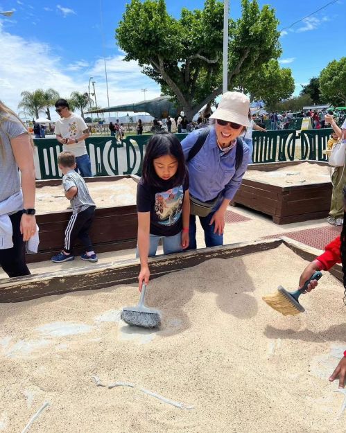Children and adults play in a sand area, using brushes and tools to explore the sand together.