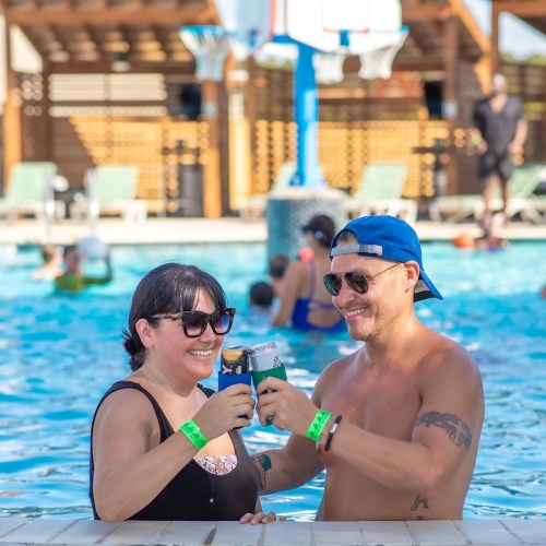 A man and woman toast with drinks in a pool, surrounded by people enjoying the water on a sunny day.