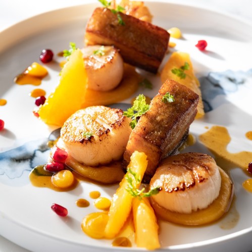A gourmet dish featuring seared scallops, crispy pork, and citrus segments, garnished with herbs and colorful sauces.
