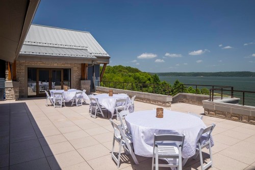 Outdoor terrace with white tables and chairs overlooking a scenic lake and green hills under a clear blue sky.