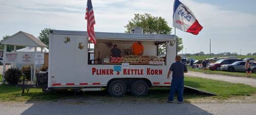A food truck selling kettle corn, with two people inside and flags flying outside, set in a rural area.