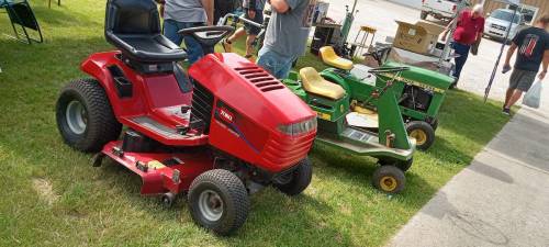 Two riding lawn mowers parked on grass: a red one on the left and a green one on the right, with people in the background.