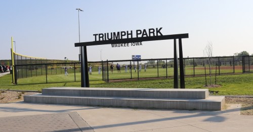 Sign for Triumph Park in Waukee, Iowa, with a baseball field and players visible in the background.