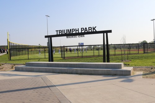 Sign for Triumph Park in Waukee, Iowa, with a baseball field and players visible in the background.