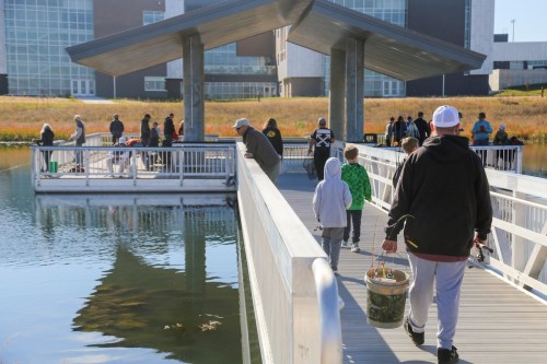 People walking on a dock by a calm waterway, with a group fishing from a nearby platform.