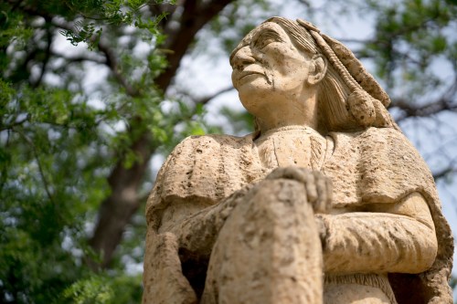 A weathered stone statue of a woman, with intricate details, surrounded by greenery.