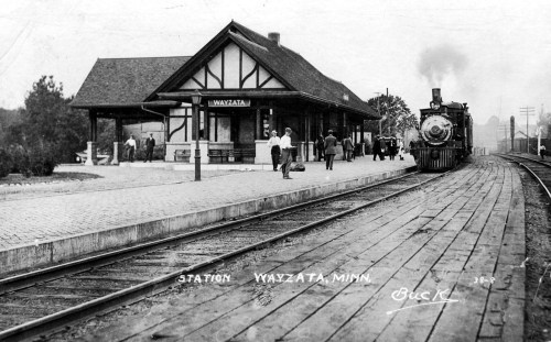 Historic black-and-white photo of Wayzata, Minnesota train station with a steam locomotive and people on the platform.