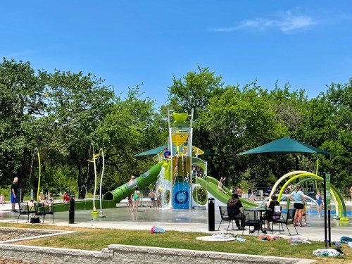 A vibrant water park with slides, splash features, and people enjoying the sun under green trees and blue skies.