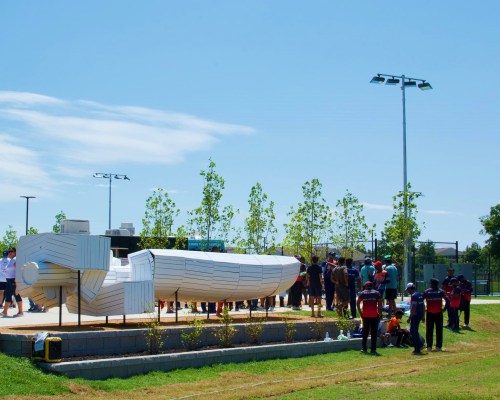 A group of people gathers around a large white sculpture resembling a boat in a sunny park setting.