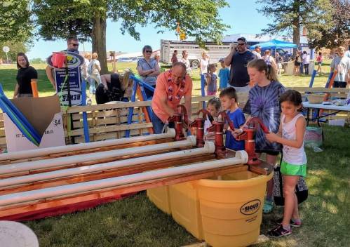 Children and adults engage with a water activity at an outdoor event, using pipes and valves in a sunny park setting.