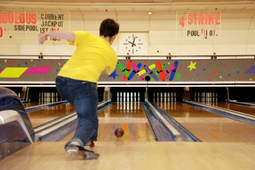 A person in a yellow shirt bowls down a lane at a bowling alley, with colorful decorations in the background.
