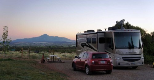 An RV parked near a mountain at sunset, with a red car and a picnic table in the foreground.