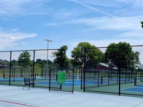 A sunny outdoor tennis court area with multiple courts, surrounded by a fence and green trees in the background.