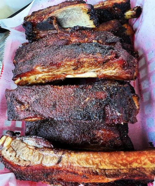 A close-up of a platter of smoked, charred beef ribs arranged on pink paper.