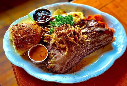 A plate of tender brisket topped with crispy onions, served with sides of barbecue sauce, bread, and vegetables.